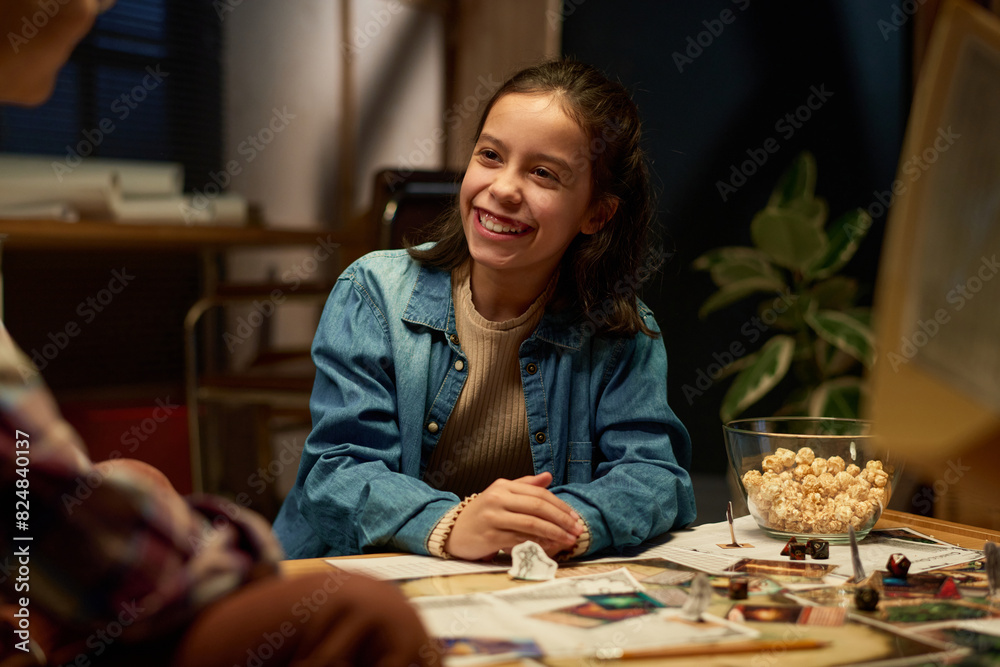 Happy brunette schoolgirl looking at her friend with toothy smile during chat or discussion of rules of popular tabletop game