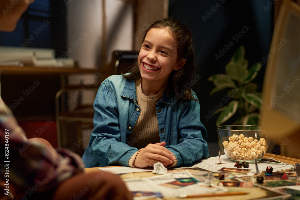 Fototapeta premium Happy brunette schoolgirl looking at her friend with toothy smile during chat or discussion of rules of popular tabletop game