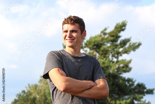 Young smiling man on the summer day. Happy and confident man with crossed arms. Candid young man outside. Nature blurred background