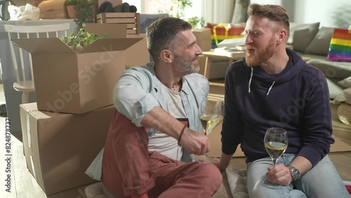 Happy young gay male couple having wine in new home after moving in