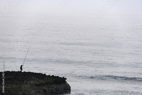 Solitary Fisherman on Rocky Coastline in Misty Conditions