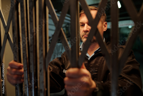 Obraz na plátně Serious mature male guard standing behind mesh and opening gate before entering