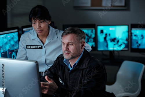 Young female and mature male security guards in uniform looking at computer screen while one of them explaining detail of video