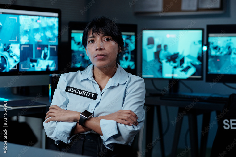 Serious young female security guard in uniform keeping arms crossed by chest and looking at camera against row of computer monitors