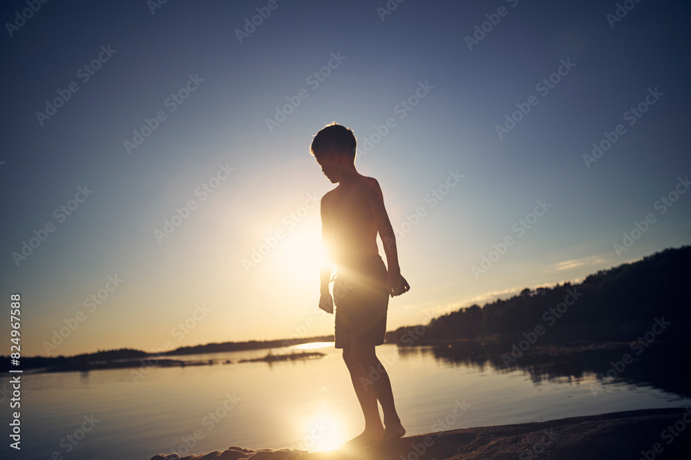 Full length of silhouette boy standing at lakeshore against sky at sunset Full length of silhouette boy standing at lakeshore against sky at sunset