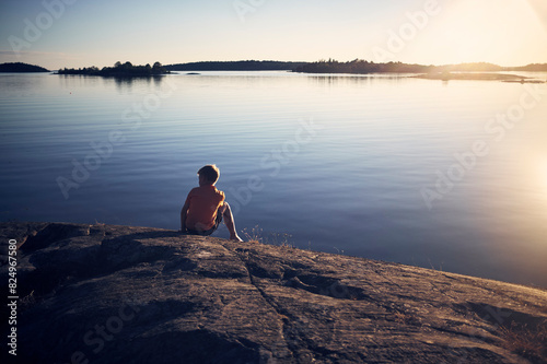 Rear view of boy sitting on rock's edge at lakeshore during sunset photo