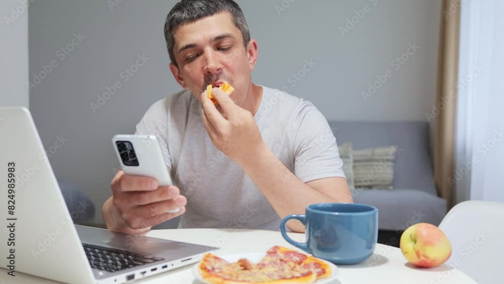 Hungry attractive young man using mobile phone while working on laptop at home feeling hungry taking break during remote job eating tasty fast food Hungry attractive young man using mobile phone while working on laptop at home feeling hungry taking break during remote job eating tasty fast food