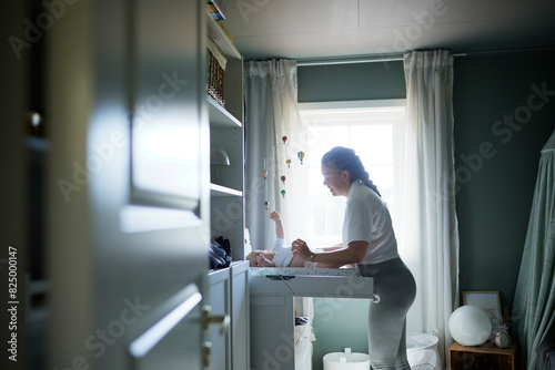 Side view of young mother changing baby's diaper while standing by window photo