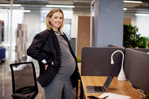 Portrait of a beautiful smiling pregnant woman working in an office with a laptop, showcasing professional and maternity balance in a modern work environment