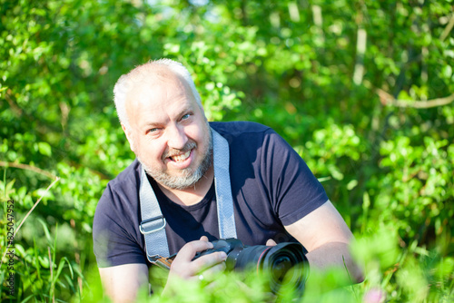 A mature man with a camera is smiling while capturing outdoor photography in nature with a greenery background © Bjorn B