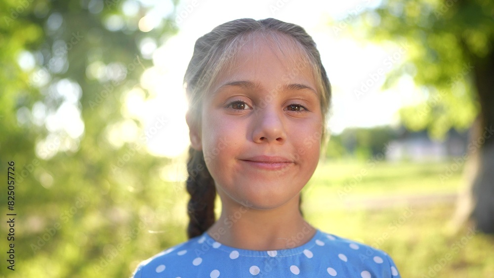 the little girl smiles. concept of a happy childhood and loving family. girl schoolgirl looks at the camera and smiles widely, face close-up, park lifestyle and sun glare in the background