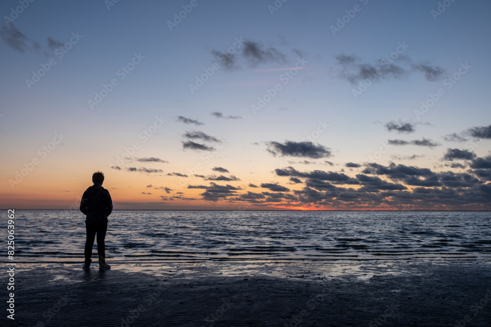 A person is on the beach, watching the sunset under a cloudy sky, with a tranquil sea spreading out before them