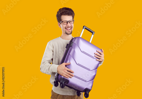 Portrait of happy young man who happily laughs holding small travel suitcase in his hands. Young Caucasian man is happy about long-awaited vacation isolated on yellow background. Travel concept.