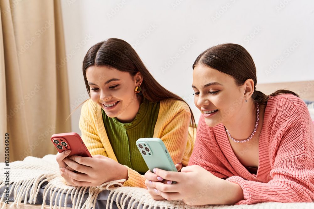 Two young women in casual clothes lay side by side on a bed, engrossed in their cell phones. Two young women in casual clothes lay side by side on a bed, engrossed in their cell phones.