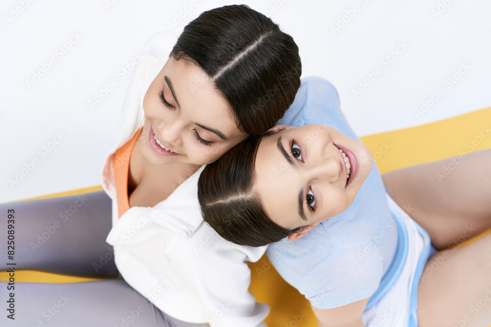 Two pretty brunette teenage girls in sportive attire sitting next to each other on a grey background in a studio. Two pretty brunette teenage girls in sportive attire sitting next to each other on a grey background in a studio.
