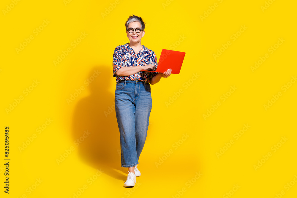 Full size photo of nice pensioner female hold netbook walking wear trendy colorful outfit isolated on yellow color background Full size photo of nice pensioner female hold netbook walking wear trendy colorful outfit isolated on yellow color background