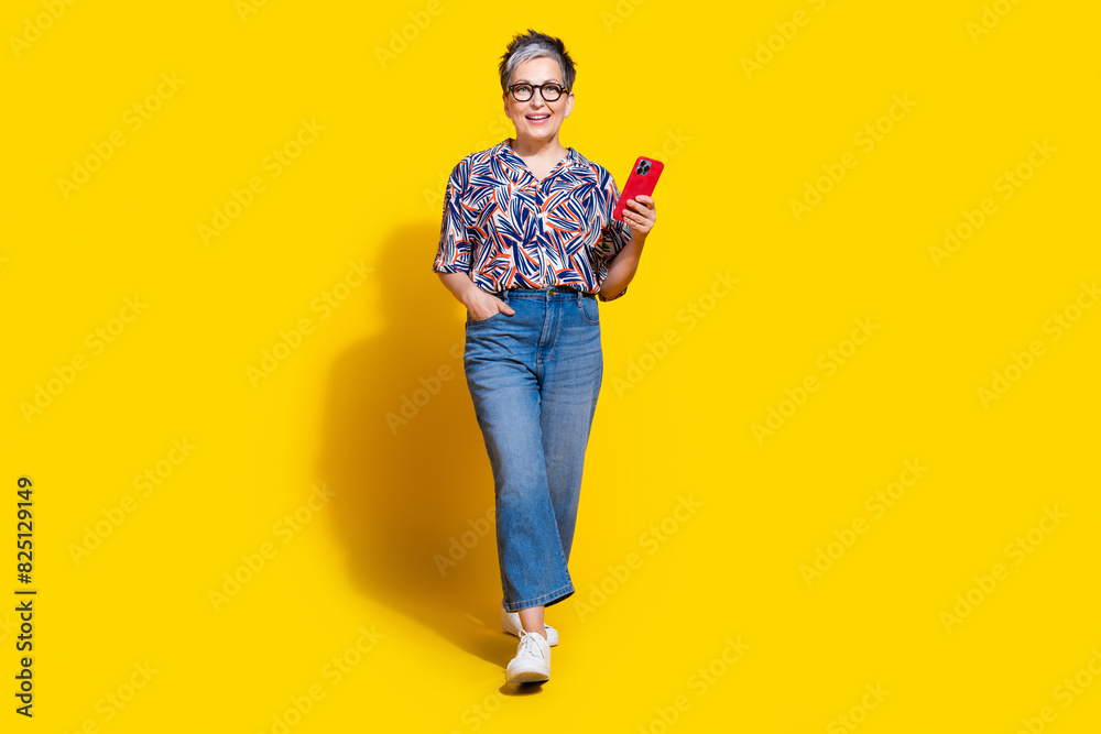 Fototapeta premium Full body photo of attractive retired woman walk hold device dressed stylish colorful clothes isolated on yellow color background