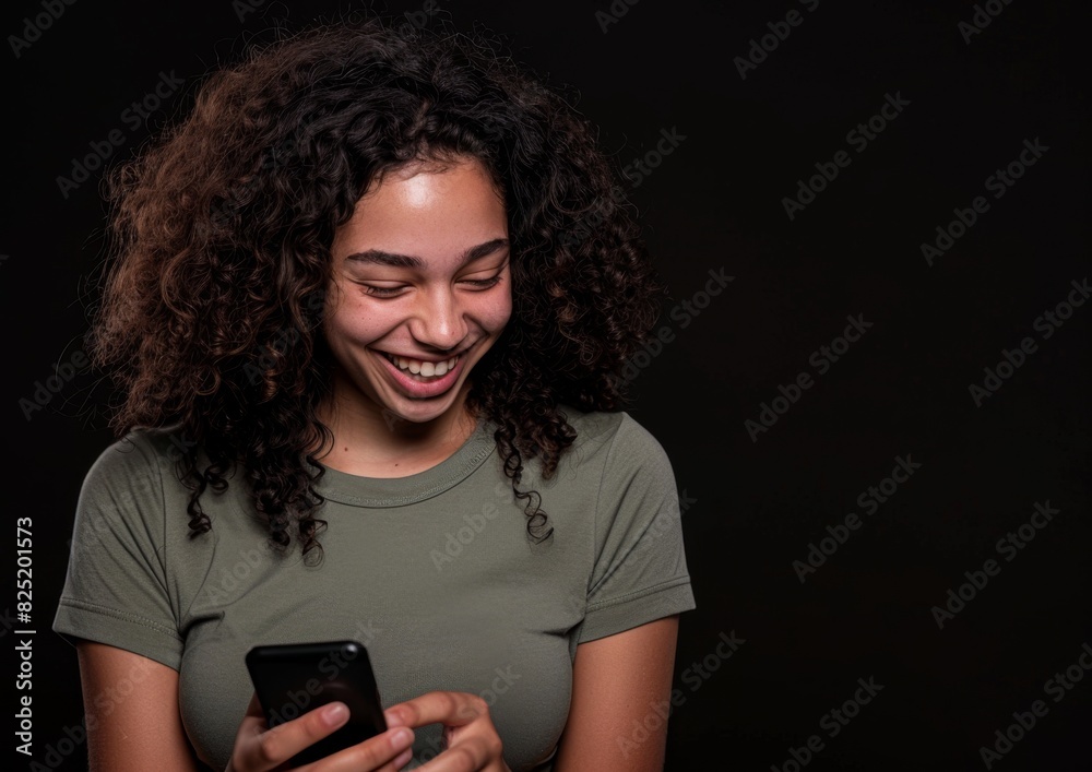 A young woman smiles as she looks at her cell phone, which has a black background, Happy woman using cell phone A young woman smiles as she looks at her cell phone, which has a black background, Happy woman using cell phone