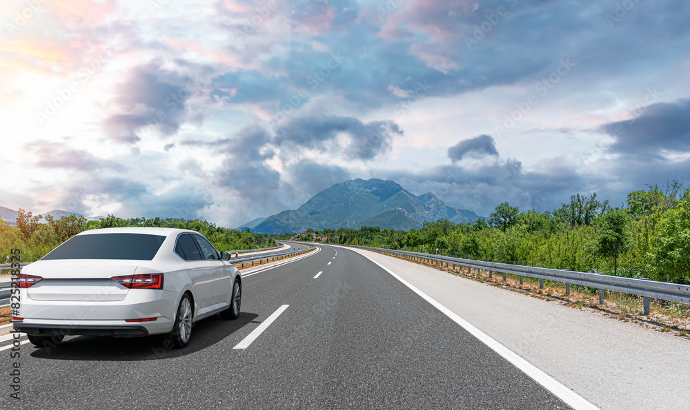 A white car drives along the highway against the backdrop of rocky mountains on a sunny day.