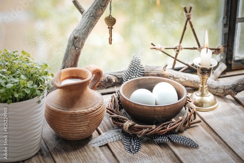 Home atmospheric spring altar with willow wreath, clay jug with milk, bowl with eggs, the first greens in the pot, candle and birds feathers on wooden surface, selected focus.