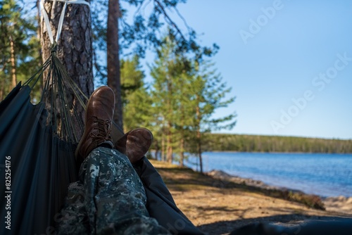 Man resting in hammock outdoors, closeup. First-person view on the legs of a man lying in a hammock overlooking a forest lake and conifers.