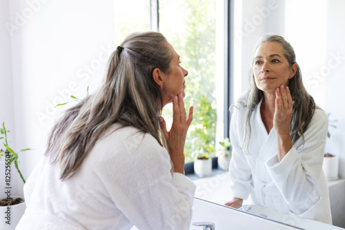 At bathroom at home, mature Caucasian woman wearing white robe, examining her face in mirror