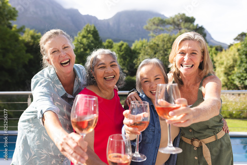 Diverse senior female friends holding wine glasses, laughing outdoors