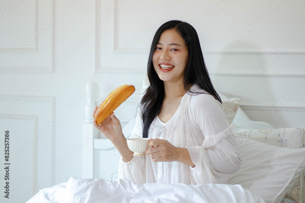 beautiful woman sitting in bed, smiling happily, holding a pastry in one hand and a cup of tea or coffee in the other. beautiful woman sitting in bed, smiling happily, holding a pastry in one hand and a cup of tea or coffee in the other.