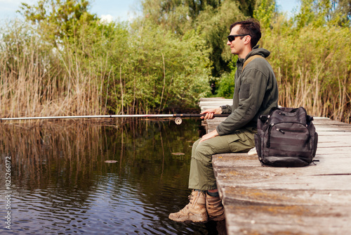 Young man fishing wearing military uniform. Guy sitting on bridge across river holding rod. Recreation