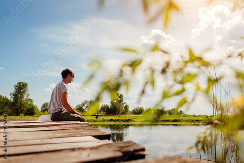 Young man fishing. Guy sitting on bridge across lake holding rod on spring day. Recreational outdoors activities