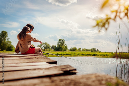 Young couple in love fishing sitting on bridge across lake holding rod on spring day. Outdoor recreational activities