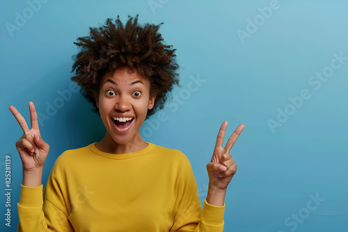 Horizontal shot pleased dark skinned female with Afro haircut, points away with both fore fingers, shows blank space for your promotion, isolated over blue background. People, advertisement concept photo