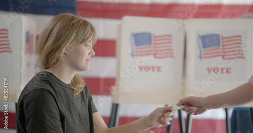 Medium shot, profile, young, red-haired woman, poll worker at voting center, checks identification for male voter. Voting booths and US flag behind