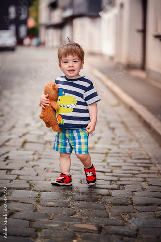Canvas Print A cheerful young boy holding a teddy bear walks on a cobblestone street in a residential area
