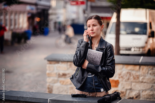 A young woman in a black leather jacket holds a notebook while talking on her phone in an urban setting. She appears focused and engaged in her conversation.