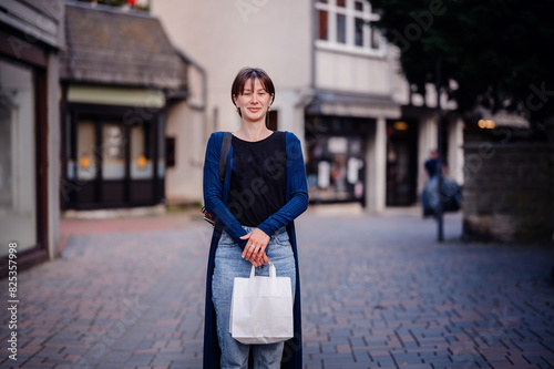 A woman with short hair smiles widely as she holds a shopping bag on a quaint town street. Dressed casually in jeans and a cardigan, she enjoys a pleasant day out.