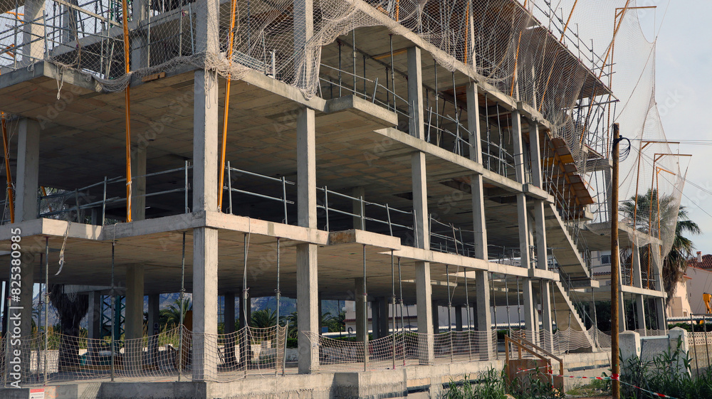 Multi-story concrete building is under construction, featuring scaffolding and safety nets. The project is located outdoors on clear land with blue sky overhead. Multi-story concrete building is under construction, featuring scaffolding and safety nets. The project is located outdoors on clear land with blue sky overhead.