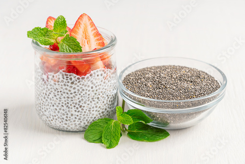 Sweet healthy chia pudding dessert made with milk decorated with topping of fresh mint leaves and sliced ripe juicy strawberries served in glass jar with bowl of seeds ingredient on white wooden table