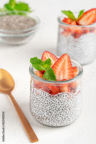 Homemade healthy chia pudding dessert made from seeds soaked in milk decorated with topping of fresh mint leaves and sliced ripe juicy strawberries served in glass bowl with spoon on white table