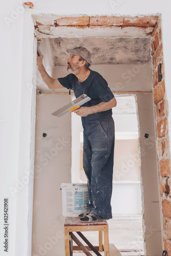 Construction worker on the construction site. wearing worker overall with wall plastering tools renovating apartment house. Plasterer renovating indoor walls and ceilings with float and plaster.  © Cristina