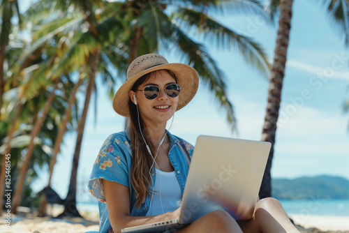 A woman wearing a hat and sunglasses working on a laptop while sitting on a tropical beach.