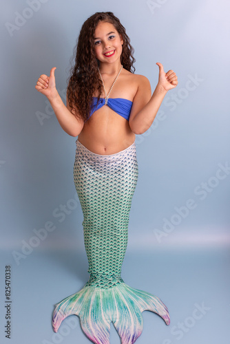 Beautiful happy girl in a mermaid costume stand on a blue background showing thumbs up.