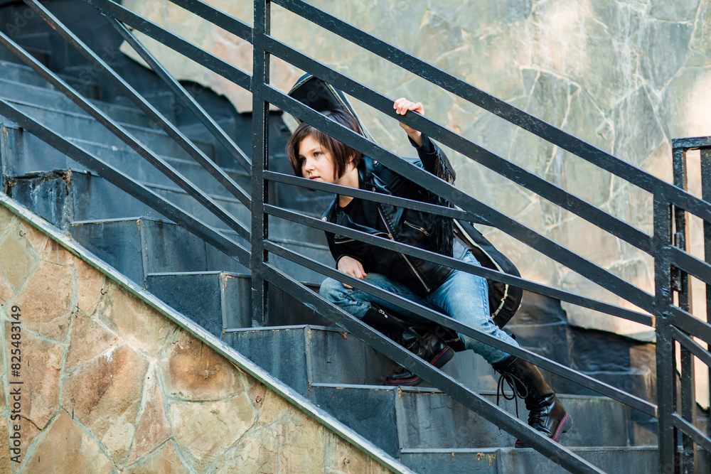 A beautiful, sweet girl standing on the stairs in jeans and a leather jacket, with an acoustic guitar in a case A beautiful, sweet girl standing on the stairs in jeans and a leather jacket, with an acoustic guitar in a case