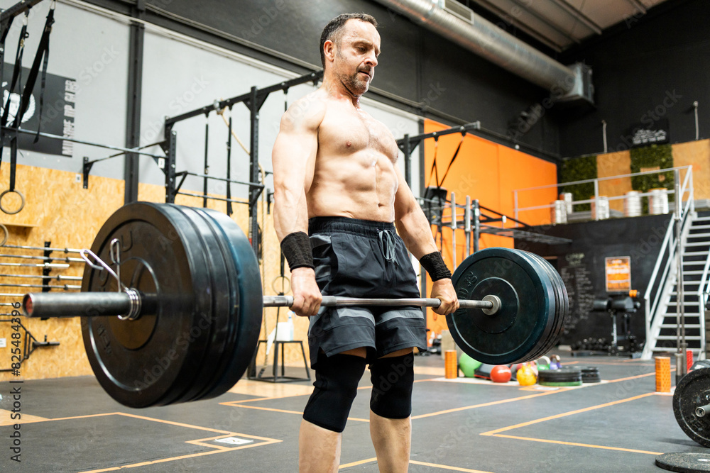 Side view of a middle-aged man without a shirt doing sports inside a gym. A 50-year-old man deadlifts to work his quads and lower back.Men between 40 and 50 years old doing weightlifting.