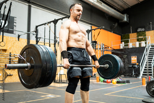 Side view of a middle-aged man without a shirt doing sports inside a gym. A 50-year-old man deadlifts to work his quads and lower back.Men between 40 and 50 years old doing weightlifting.