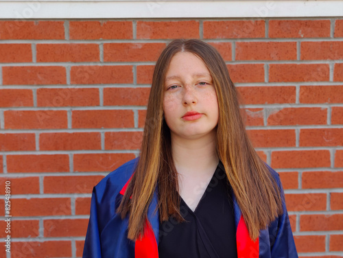 The image shows a young girl with long, straight hair standing in front of a red brick wall. She is wearing a high school graduation dress. The girl is not happy and looks directly at the camera.
