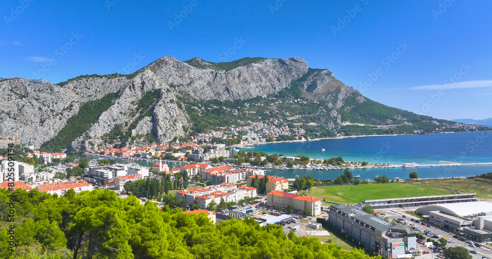 Fototapeta premium AERIAL: Scenic view of a mountain range towers above the coastal town of Omis.