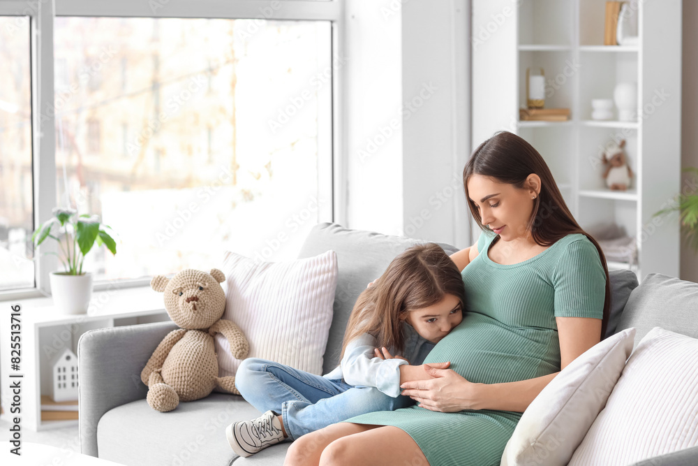 Little girl kissing belly of her pregnant mother on sofa at home