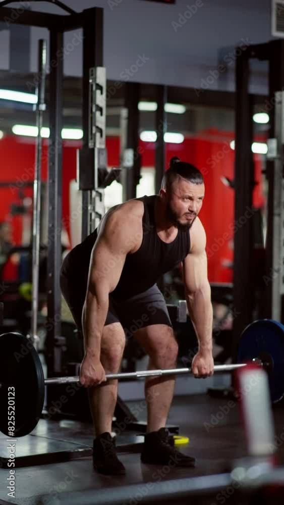 Muscular man with a beard and shaved head, wearing a black tank top and shorts, performing deadlifts with a barbell in a gym with red walls and exercise equipment Muscular man with a beard and shaved head, wearing a black tank top and shorts, performing deadlifts with a barbell in a gym with red walls and exercise equipment