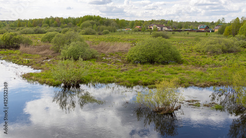  A tranquil wetland scene with reflections of lush greenery and trees in the calm water, bordered by a rural community with houses and overgrown vegetation.